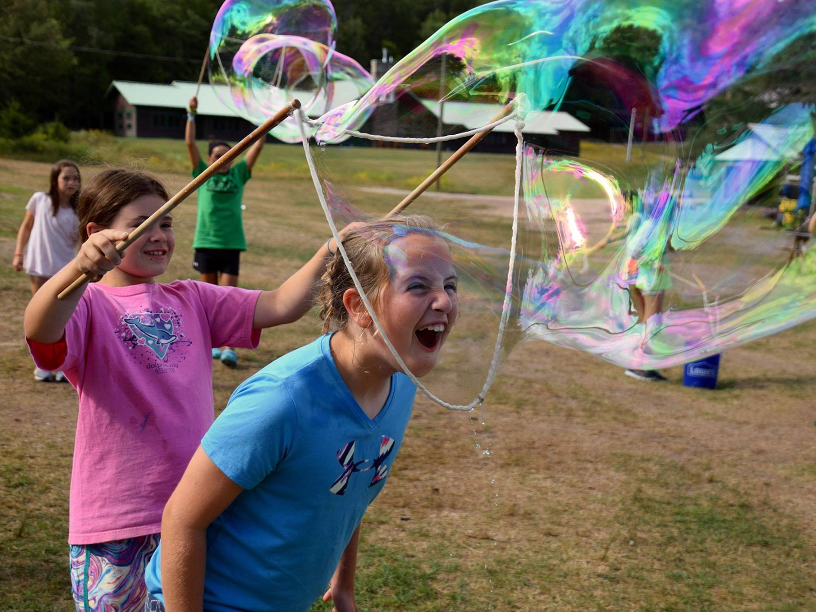 A girl laughs inside a giant rainbow bubble while her friend holds the wooden bubble sticks around her head