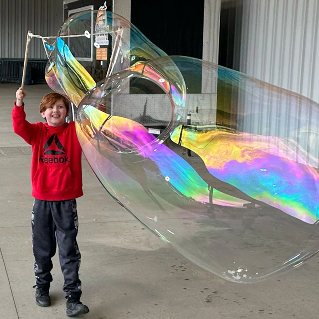 Boy in a red sweatshirt holding wooden giant bubble sticks beside a large rainbow bubble