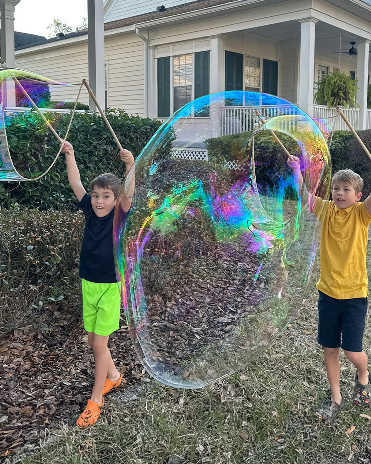 Two boys holding wooden bubble sticks while making a huge iridescent bubble