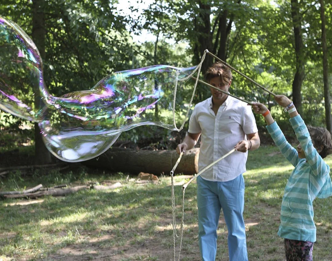 Father and daughter holding wooden giant bubble sticks beside a colorful soap film