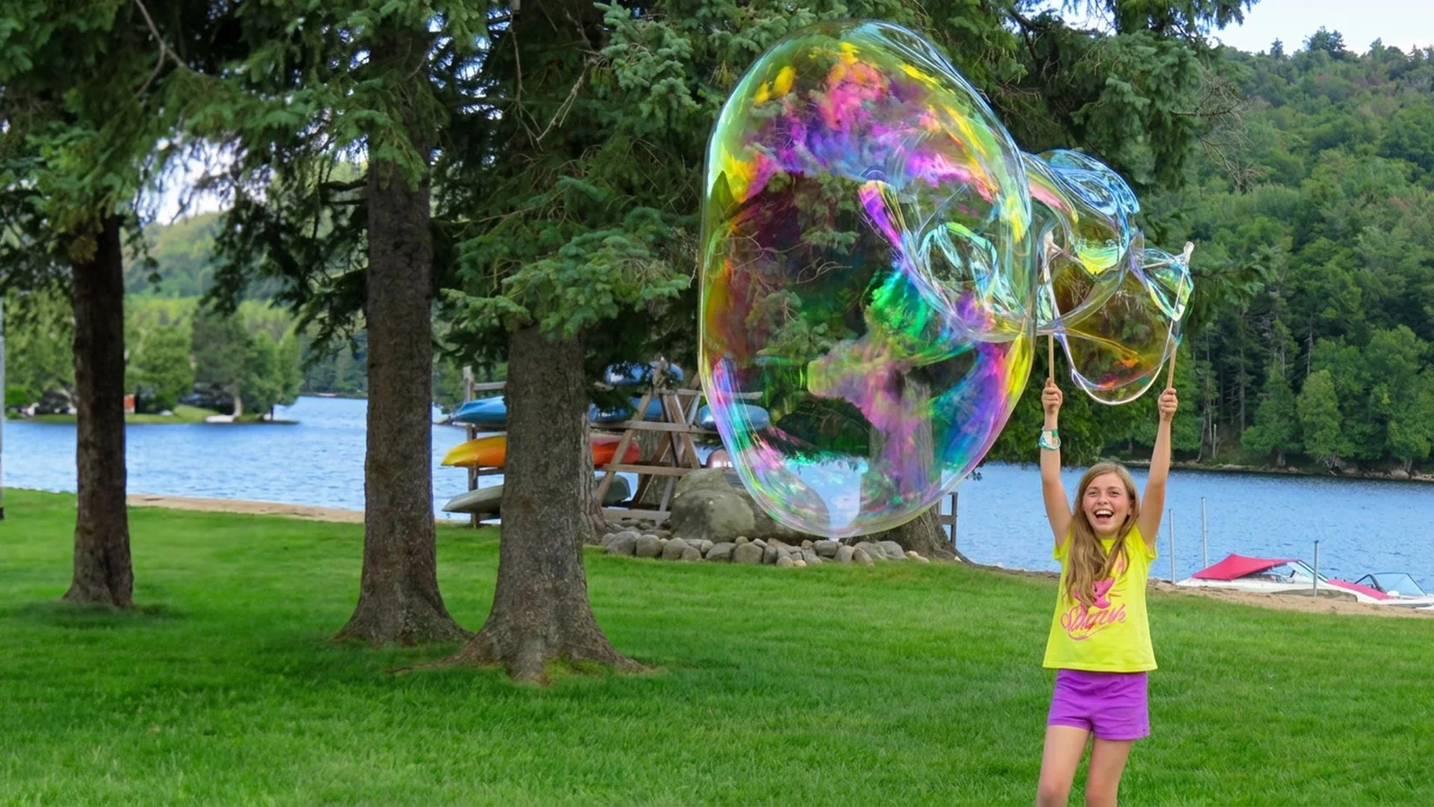 Girl raising a wooden giant bubble maker beneath a large rainbow bubble by a lake